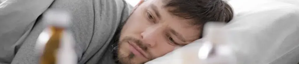A weary young man lying in bed, gazing at a bottle of pills within reach, capturing the struggle of attempting sobriety without professional help.