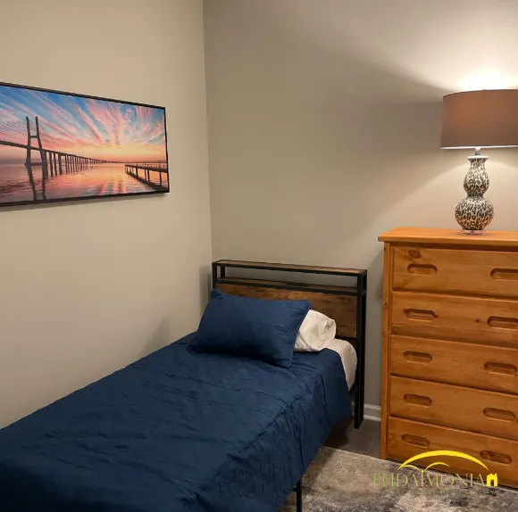 A private bedroom with a single platform bed, dresser and bedside lamp in a Houston men’s sober living home