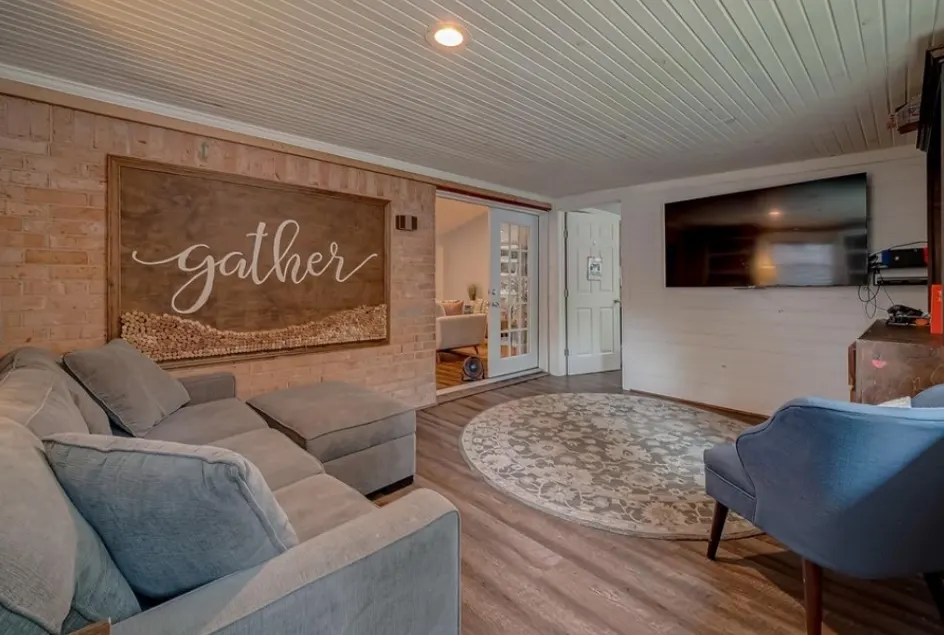 A Houston lounge with a sectional sofa, a wall-mounted television and a reclaimed-wood sign reading Gather