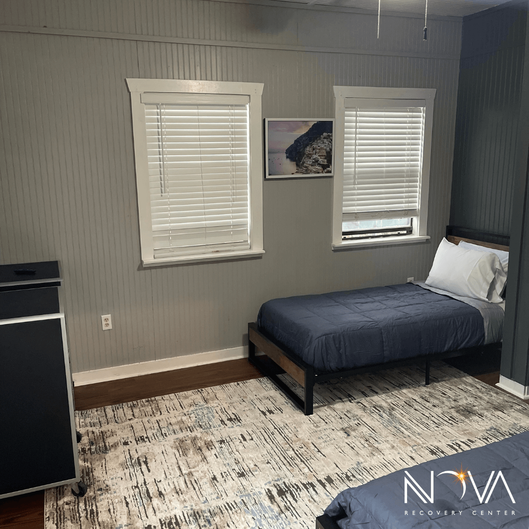Twin bed in a gray‑walled residential room with two windows and an area rug at Nova Recovery Center in Austin.
